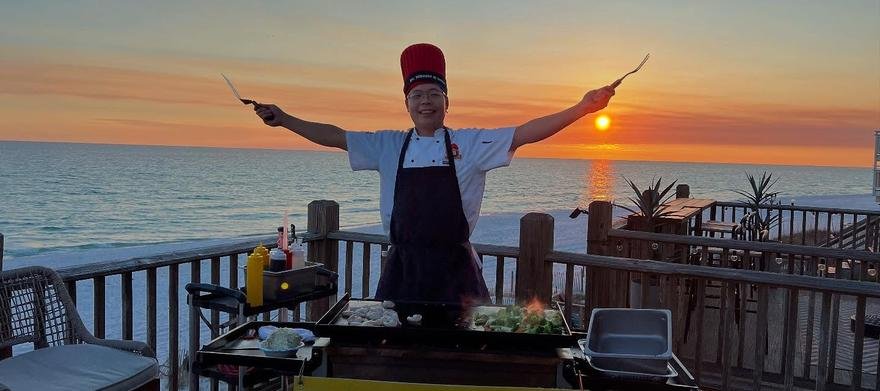 Hibachi chef raising his spatulas at sunset on a waterfront deck